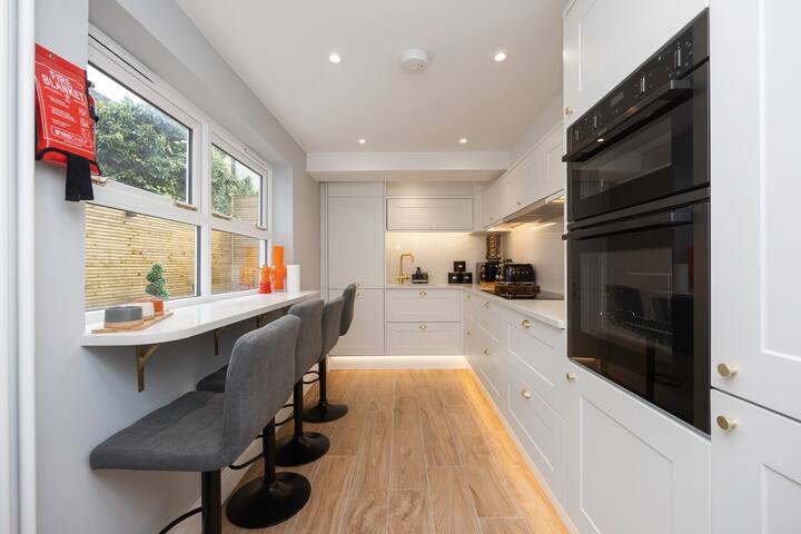 A modern kitchen with light gray cabinetry, white countertops, and light wood-look flooring. It features integrated double ovens, a sink with a gold faucet, and a breakfast bar with three gray bar stools by a window. Under-cabinet lighting illuminates the space.