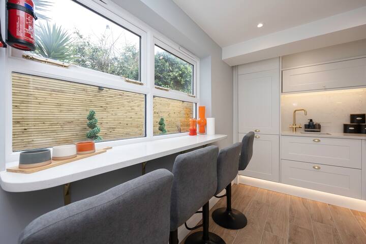 A bright kitchen bar area with a long white countertop and three gray bar stools, positioned beneath a wide window overlooking a wooden fence, with light gray cabinets and gold fixtures visible on the right.
