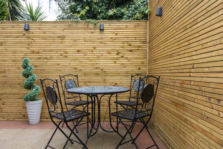 An outdoor patio area enclosed by light wooden slatted fencing, featuring a black wrought iron patio table with four matching chairs. A tall green spiral topiary plant in a white pot adds a decorative touch.