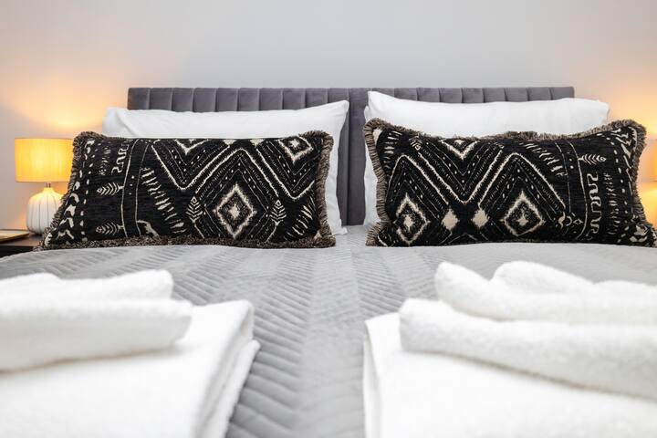 A close-up of a bed with a grey upholstered headboard, adorned with two striking black and white patterned throw pillows and crisp white sheets, alongside a grey textured duvet.