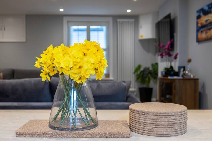 A bright living space with light gray walls and light wood flooring, featuring a large vase of yellow daffodils on a light wooden table. In the background, a gray sofa and a wall-mounted TV are visible, suggesting a living room area.