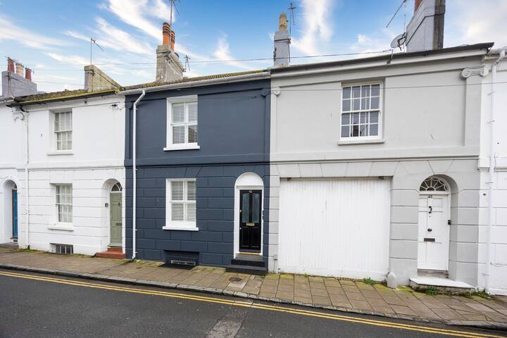 The front exterior of a terraced house with a dark grey facade, white window frames, and a black front door, situated on a street with other similar houses under a blue sky.