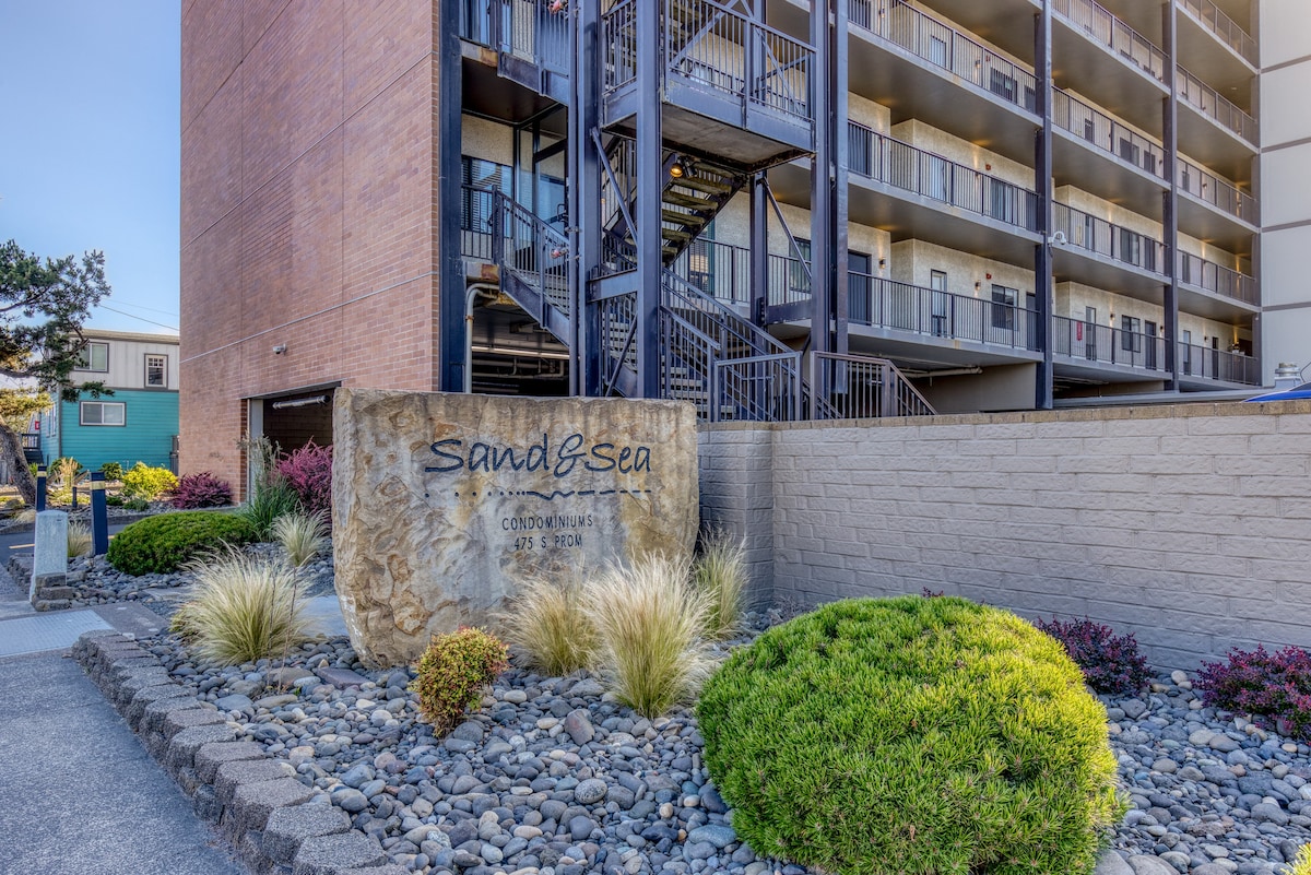 The front exterior of the Sand & Sea Condominiums, featuring a prominent stone sign, well-maintained landscaping with rocks and shrubs, and a brick facade building in the background.