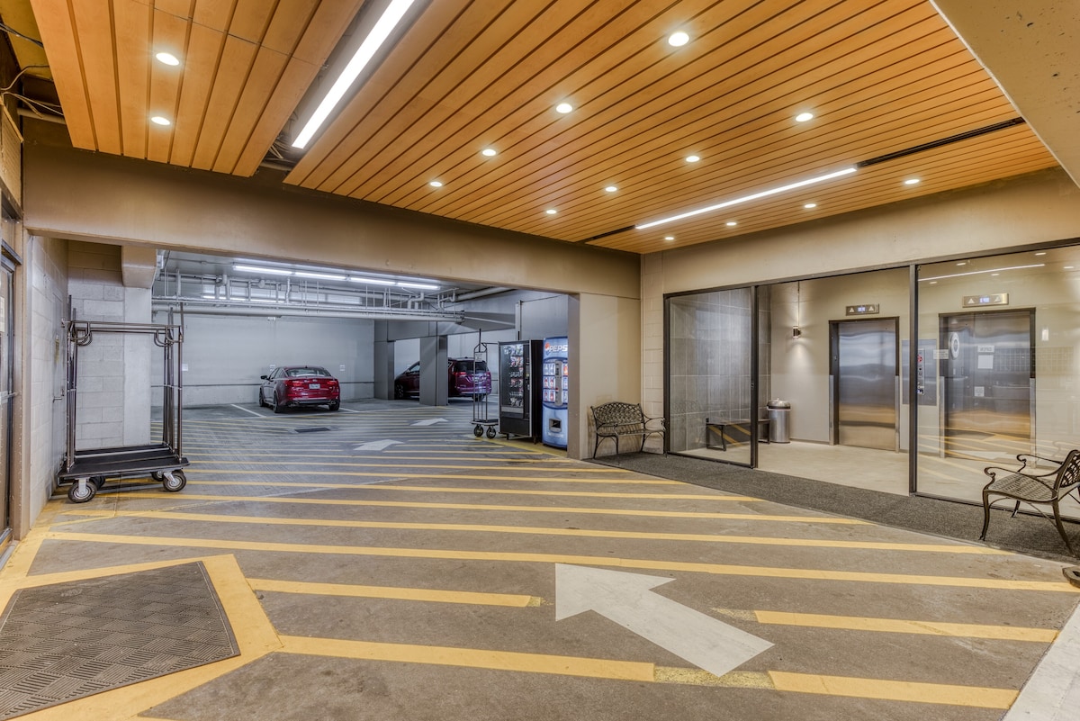 A well-lit interior of a parking garage with multiple parked cars, a vending machine, and a clear entrance area leading to the building's elevators.