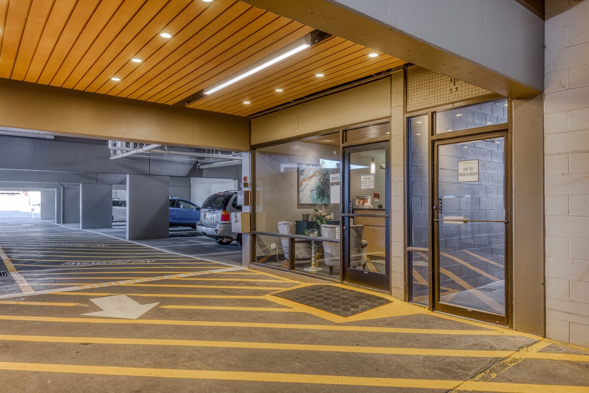 Ground-level view of the building's exterior, featuring a covered parking area with yellow striped markings and an entrance with glass doors. A sign on the right door indicates "EXIT TO PARK & BEACH".