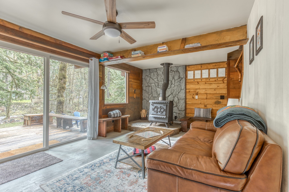 Cozy living room with a brown leather sofa, a wood-burning stove against a stone wall, and a large sliding glass door opening to an outdoor deck with forest views.