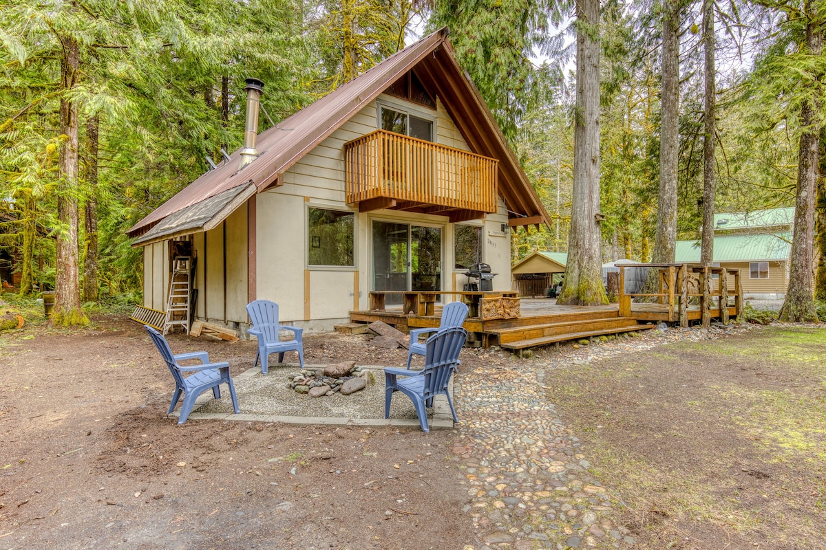 A-frame cabin with a metal roof and wooden balcony, featuring a stone-paved path leading to a fire pit area with blue Adirondack chairs, all nestled among tall evergreen trees.