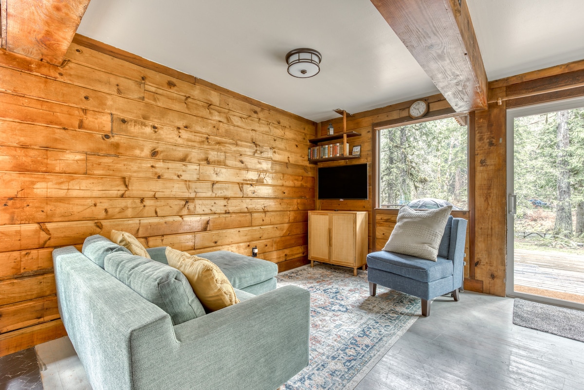 Cozy living room with a light blue sectional sofa and a matching armchair on a patterned rug, featuring a wood-paneled accent wall and a large window overlooking the forest.