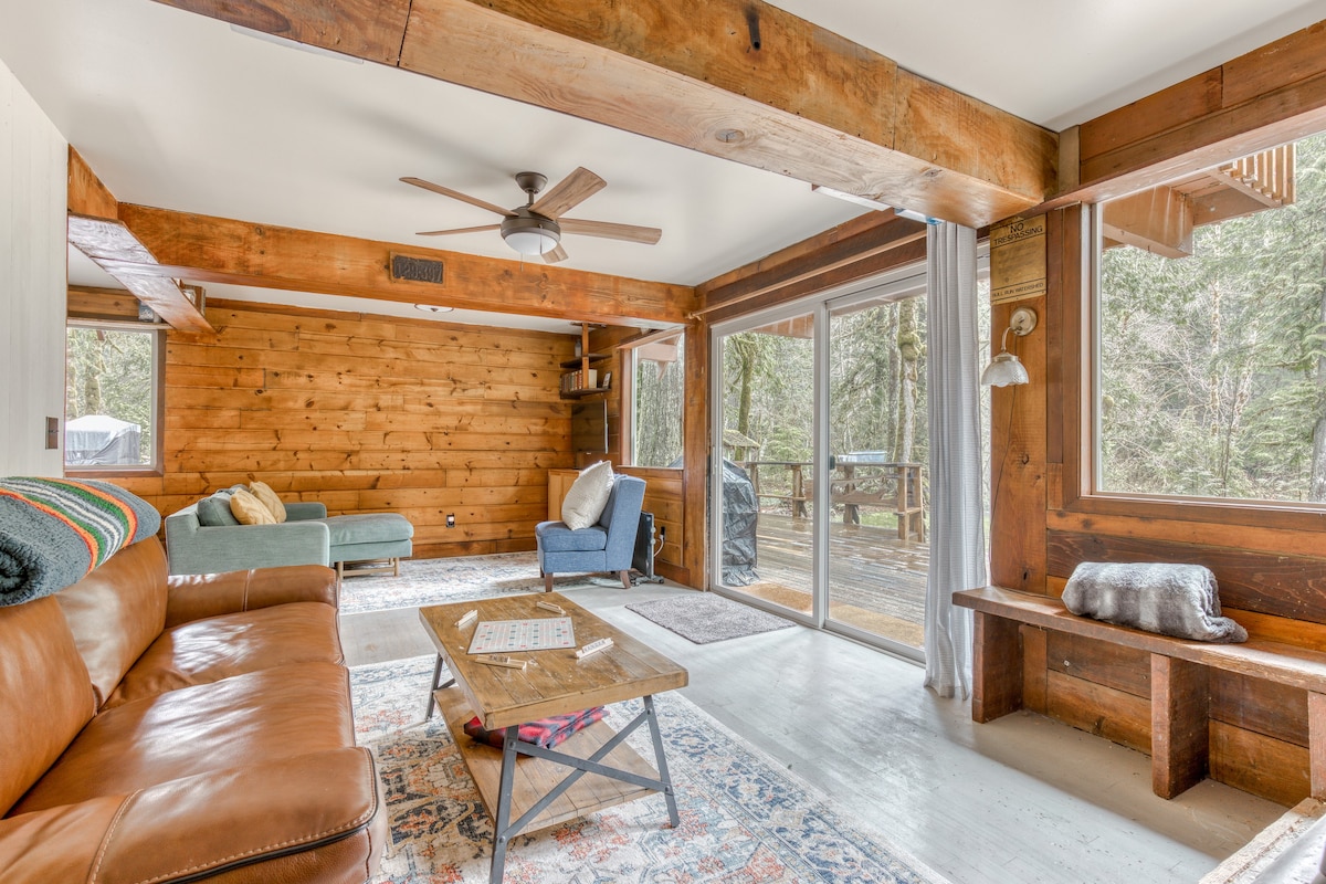 Spacious living room with a brown leather sofa, a light blue armchair, and a green chaise lounge, featuring extensive wood paneling, exposed beams, and large windows offering forest views.