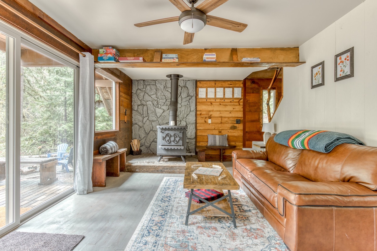 Warm living room centered around a black wood-burning stove with a stone backdrop, a comfortable brown leather sofa, and a rustic coffee table, with a sliding glass door to the outside.