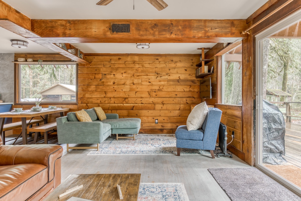 Bright living area with a light blue sectional sofa and armchair on a patterned rug, featuring a wood-paneled wall and a glass sliding door leading to an outdoor deck with a grill.