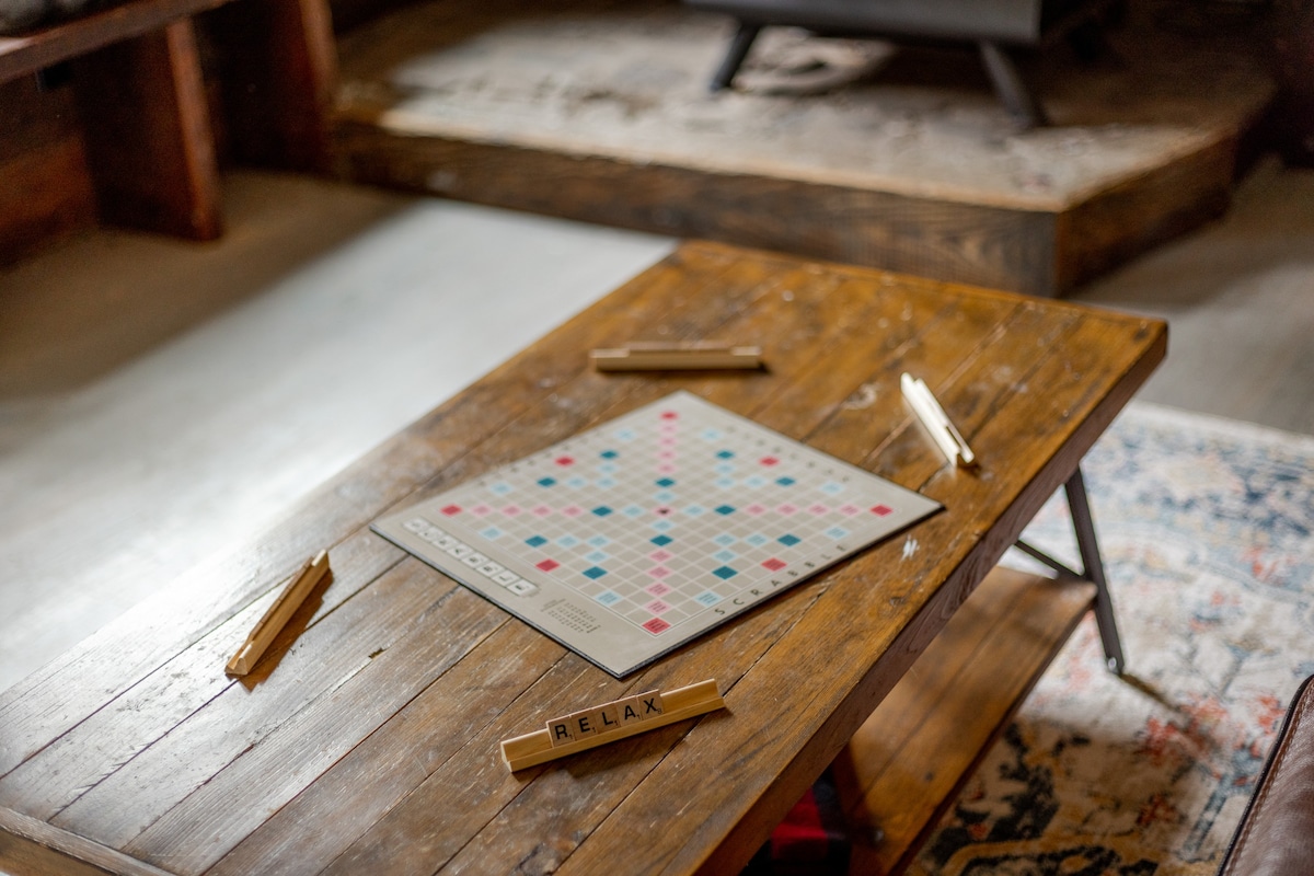 A rustic wooden coffee table with a Scrabble board and letter tiles, including 'RELAX', sits on a patterned rug in a cozy living space with a fireplace in the background.
