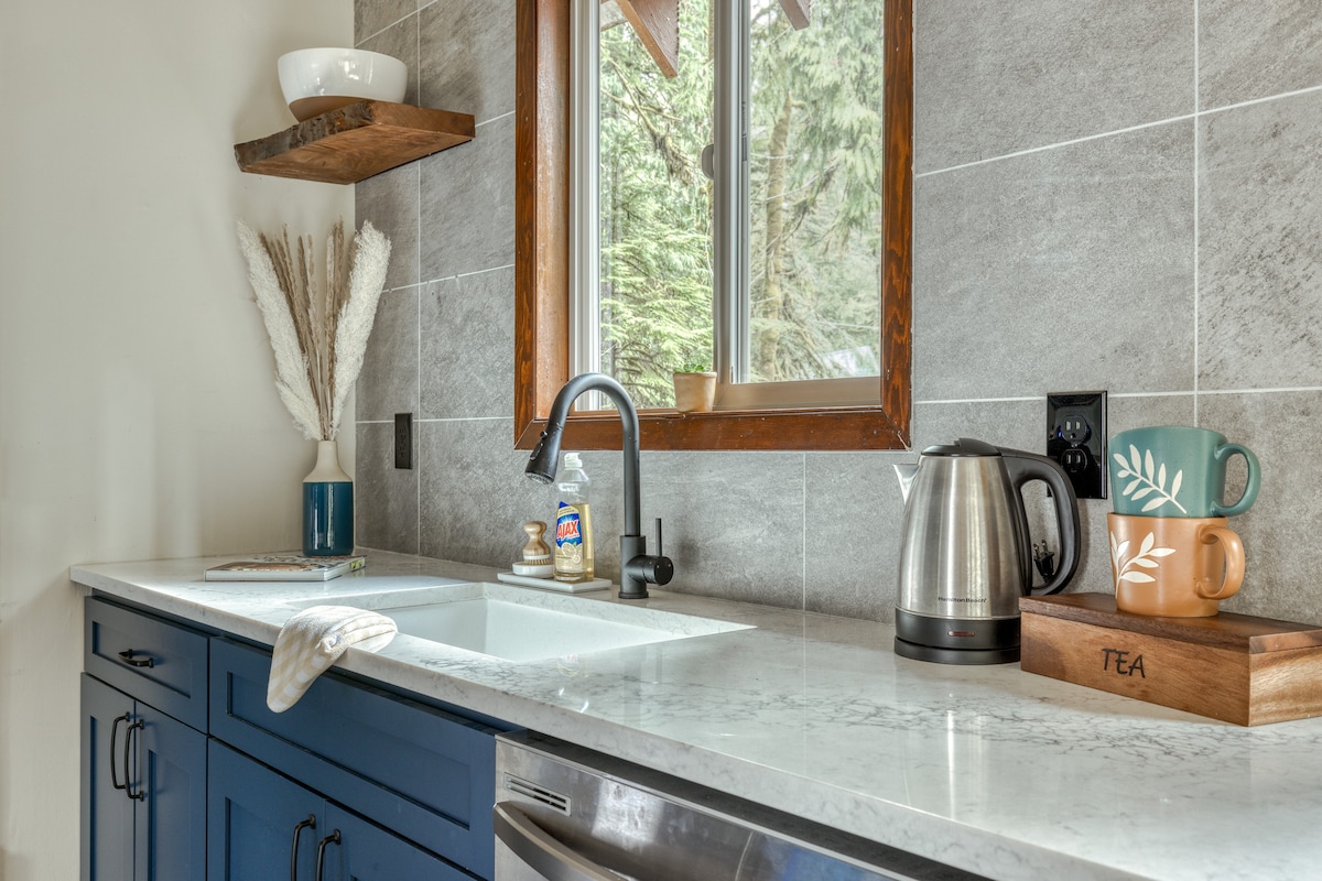 Close-up of a kitchen sink area with a black gooseneck faucet, white countertop, stainless steel kettle, and decorative mugs, set against a gray tiled backsplash and a wooden-framed window.