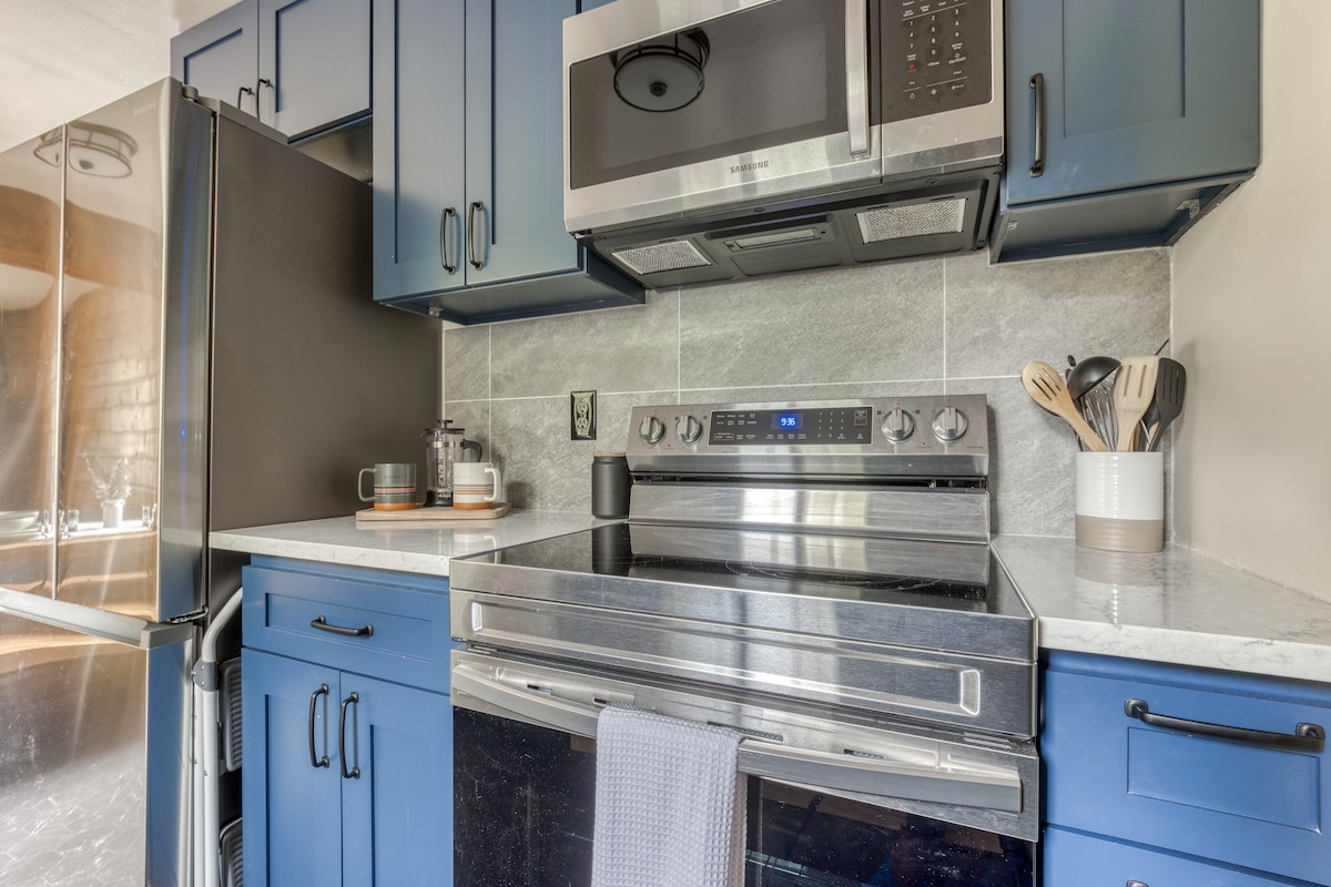 Modern kitchen with blue shaker-style cabinets, stainless steel appliances including a Samsung microwave and electric range, and light gray subway tile backsplash.
