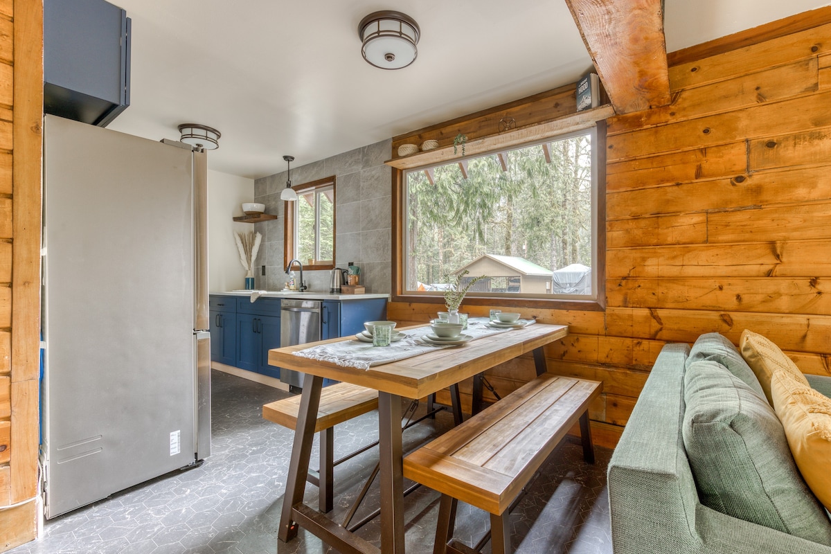 Open-concept dining area with a rustic wooden table and benches, adjacent to a kitchen with blue cabinets and a stainless steel refrigerator, all set against a wood-paneled wall.