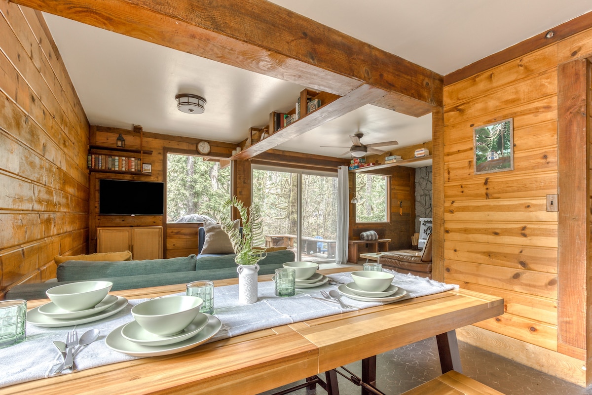 Rustic dining area with a long wooden table set with light green dishes and benches, featuring wood paneling, exposed wooden beams, and an open view into the adjacent living room.