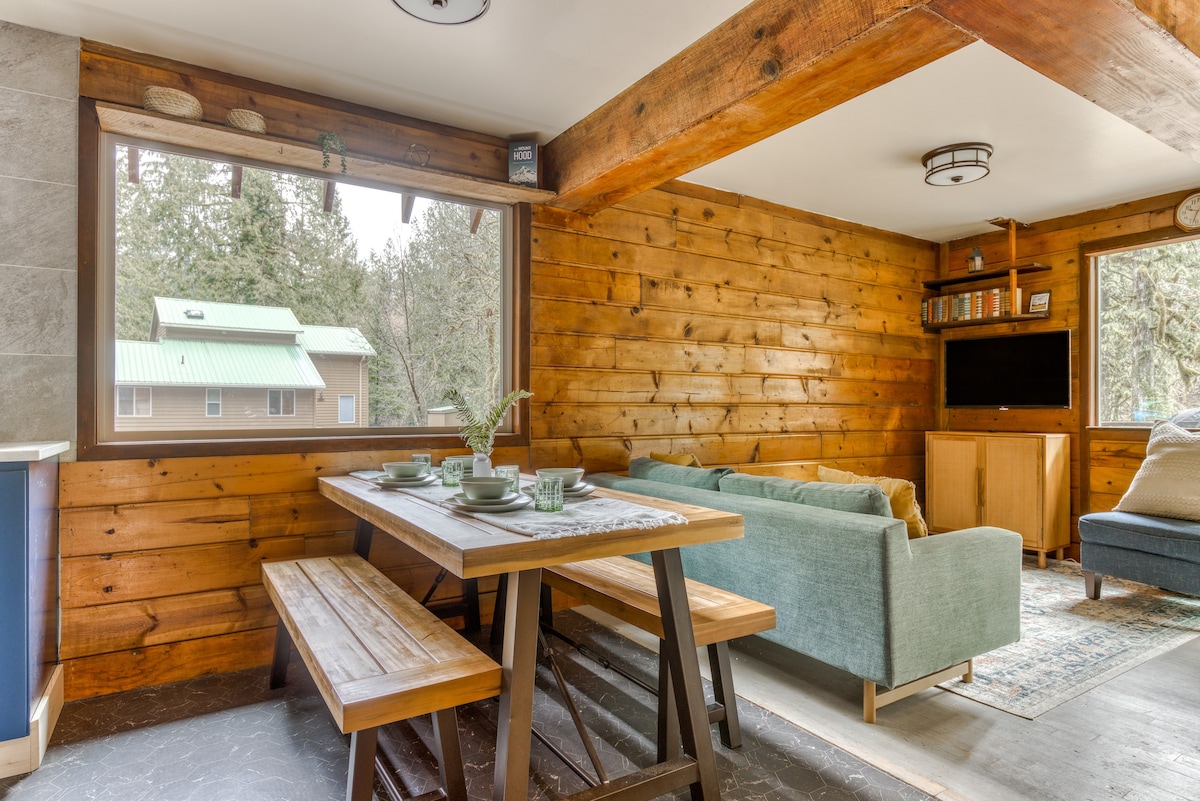 Cozy dining area featuring a long wooden table with benches, leading into a living space with a light blue sofa and a TV, all surrounded by warm wood-paneled walls and large windows.