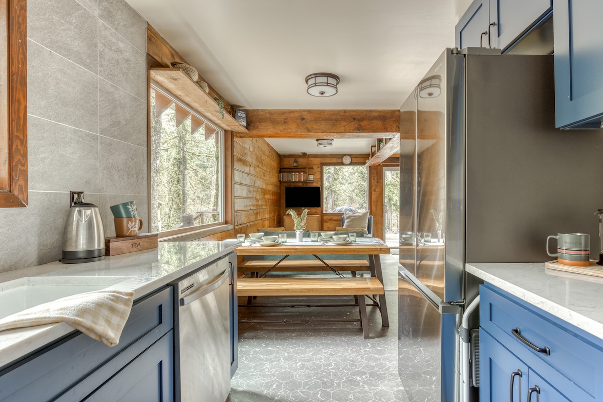 View from the kitchen into the dining area, showcasing blue cabinets and a stainless steel refrigerator on the left, leading to a wooden dining table with benches and large windows overlooking the outdoors.