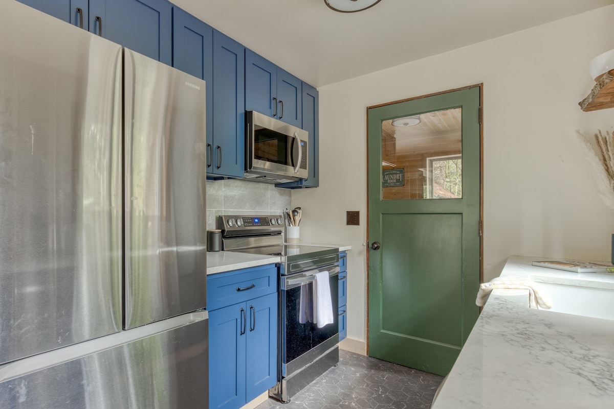 View of the kitchen showing a stainless steel refrigerator, blue upper and lower cabinets, and a stainless steel microwave and electric range. A green half-glass door leads to another room, and the floor is dark hexagonal tile.