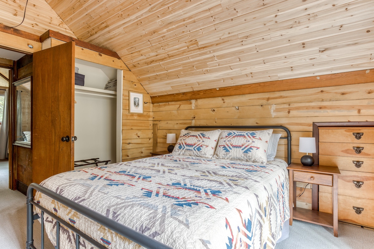 Cozy bedroom with a metal-frame queen bed featuring a colorful patterned quilt. The walls and sloped ceiling are clad in light-toned wood planks, and a wooden dresser and nightstand are visible.