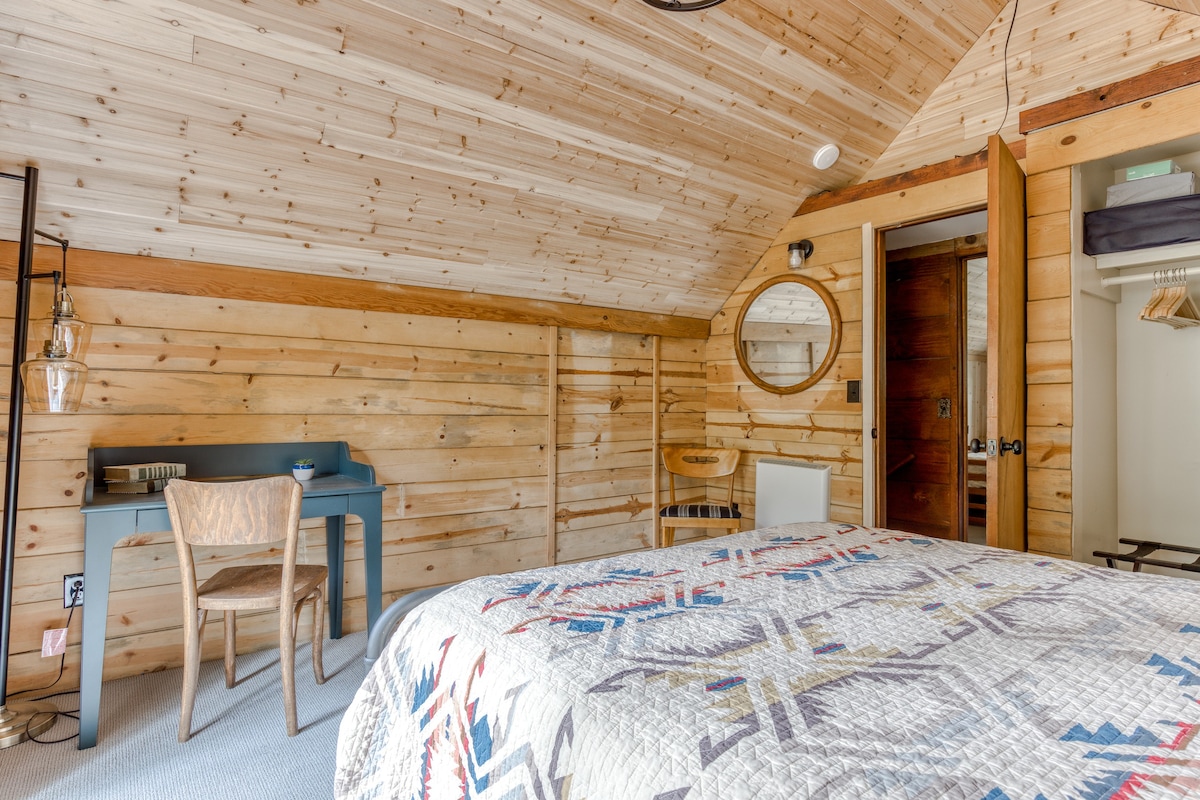 Another view of the bedroom, showing the wood-paneled walls and ceiling, a light blue desk with a wooden chair, and a round mirror above the bed. The room has a cozy, rustic cabin feel.