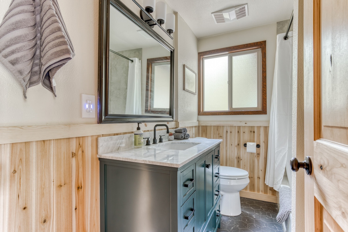 Wider view of the bathroom, featuring a dark vanity with a marble top, wood-paneled lower walls, and a toilet. A shower/tub combo with a white curtain is visible on the right, and the floor is dark hexagonal tile.
