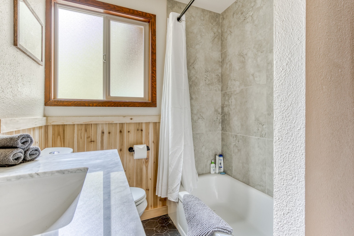Close-up of the shower/tub combo in the bathroom, featuring gray tiled walls and a white shower curtain. A window with frosted glass and a wooden frame is above the tub, providing natural light.