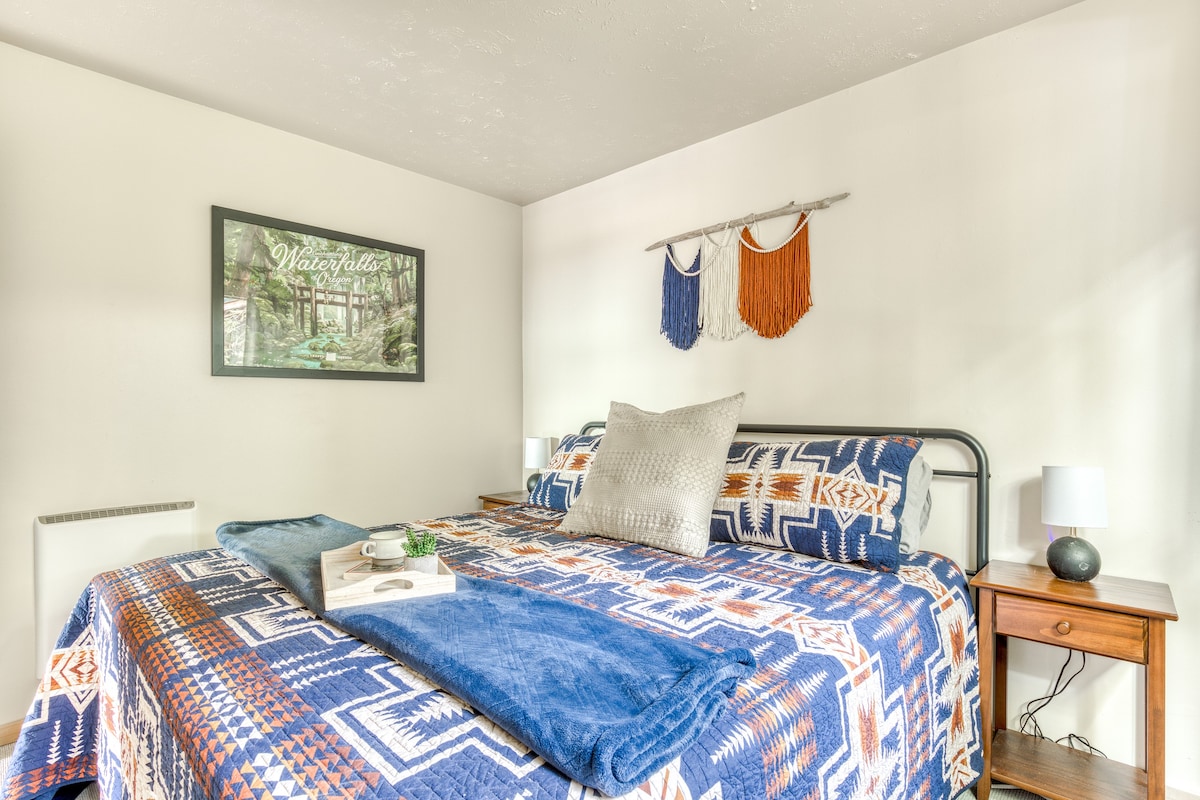A cozy bedroom featuring a large bed with a blue, orange, and white geometric patterned quilt, white walls, and a framed nature print above the bed. A macrame wall hanging adds a bohemian touch.