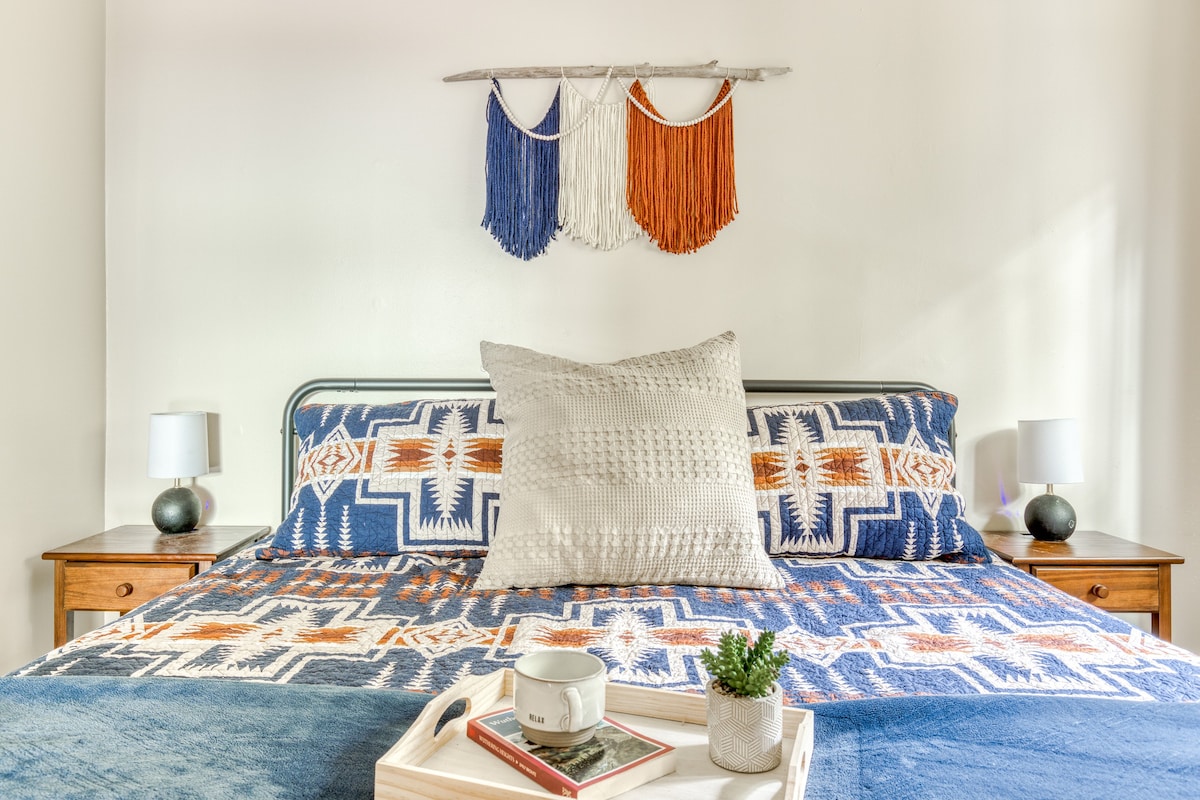 A close-up view of the bed in bedroom-1, highlighting the blue, orange, and white geometric patterned quilt, a decorative pillow, and a wooden tray with a "RELAX" mug and a small potted plant.