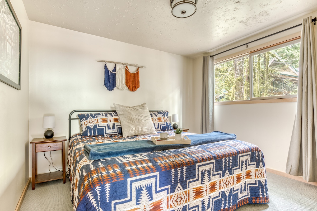 A bright bedroom with a large bed featuring a blue, orange, and white patterned quilt, flanked by wooden nightstands with small lamps. A window with light gray curtains offers a view of outdoor greenery.