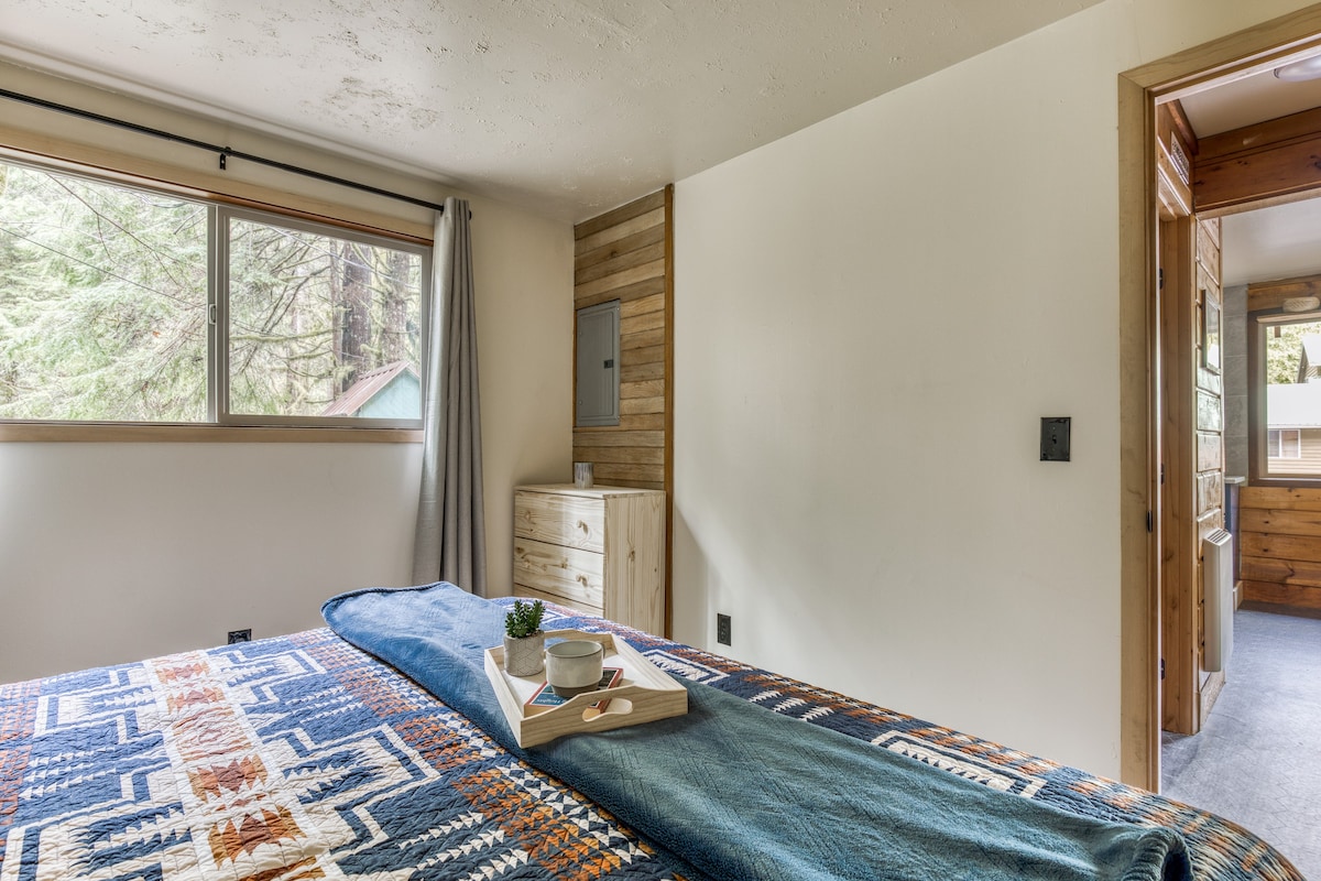 An alternate view of bedroom-1, showcasing the bed with its patterned quilt, a large window overlooking trees, and a light wood dresser against a vertical wood-paneled accent wall.