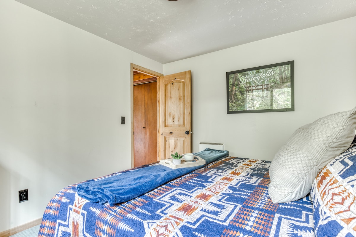 A view of bedroom-1 showing the bed with its patterned quilt, a framed nature print on the wall, and an open wooden door leading to another part of the cabin.