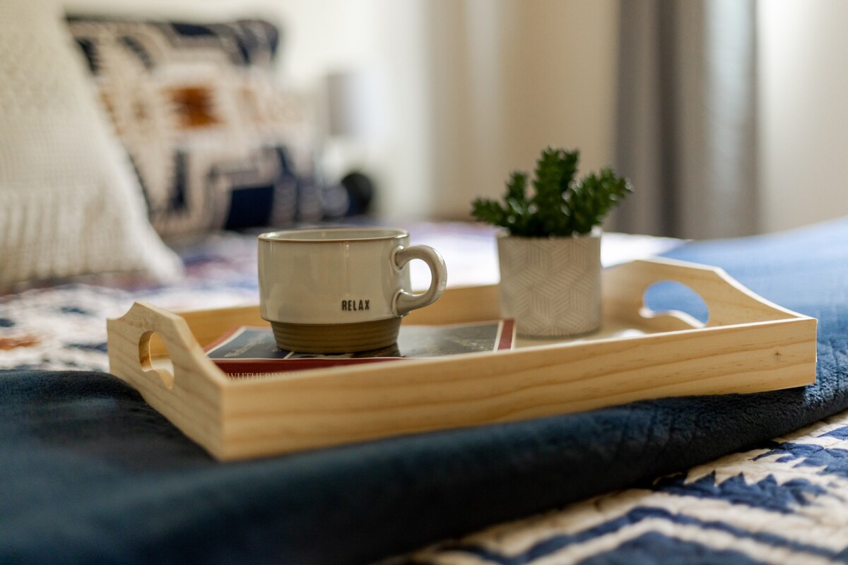 A detailed close-up of a light wooden tray placed on the bed, holding a cream-colored mug with "RELAX" inscribed and a small green potted plant.