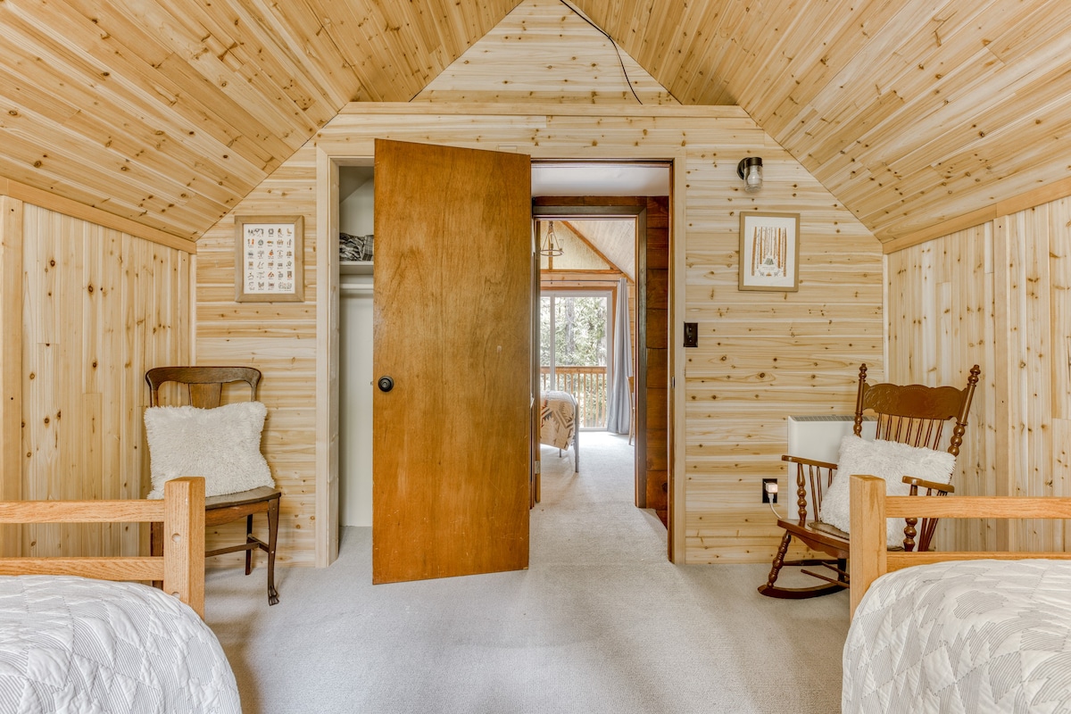 A rustic bedroom with light wood paneling on the walls and sloped ceiling, featuring two twin beds with white quilts and a classic wooden rocking chair with a fluffy white pillow.