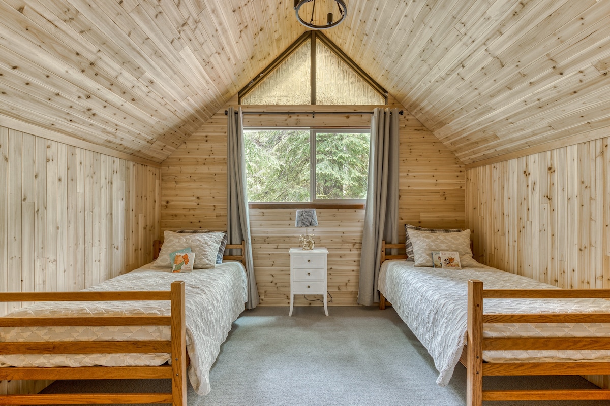 A symmetrical view of bedroom-2, featuring two identical twin beds with white patterned quilts, separated by a white nightstand, all within a room with light wood-paneled walls and a high, sloped ceiling.