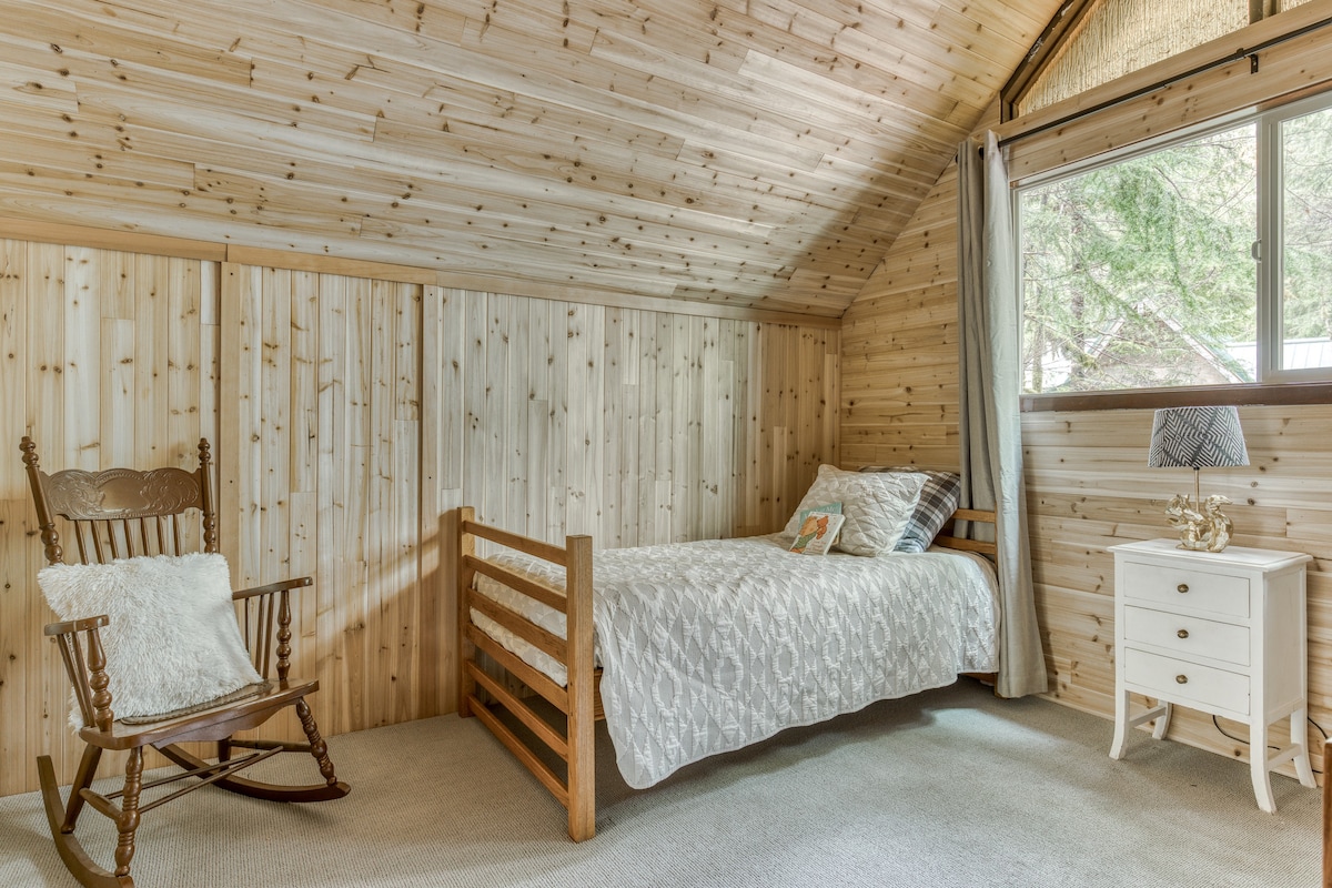 A cozy corner of bedroom-2, showcasing a twin bed with a white patterned quilt, a white nightstand with drawers, and a decorative lamp, all against light wood-paneled walls and a sloped ceiling.