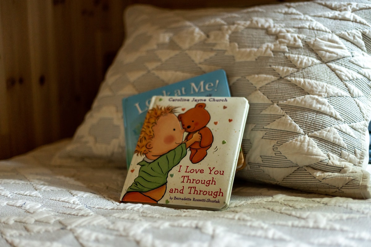 A close-up view of a bed with a white quilted comforter and a patterned pillow, featuring two children's books, one titled 'I Love You Through and Through' and another partially visible.
