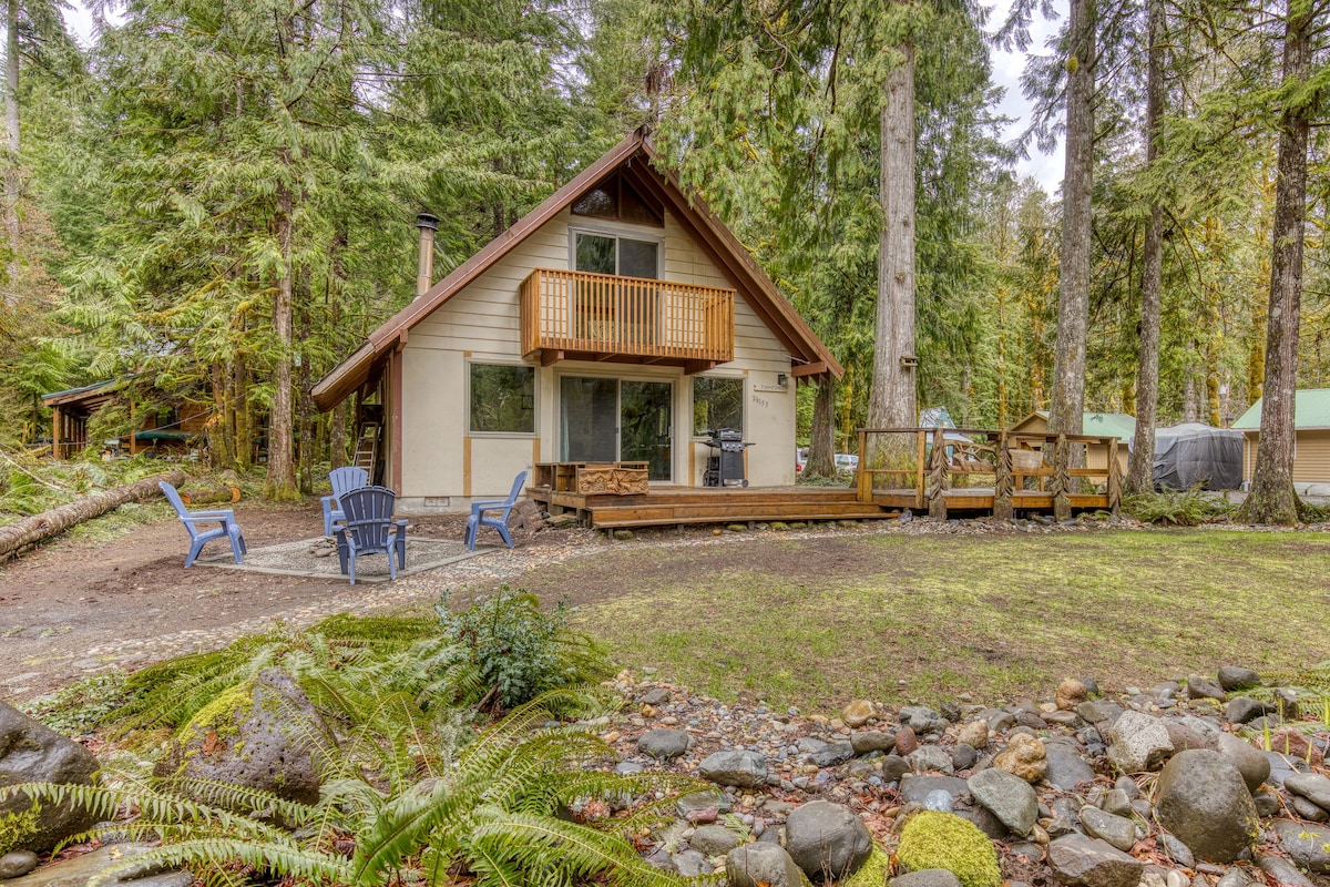 A charming A-frame cabin with light-colored siding and a wooden balcony, nestled among tall evergreen trees, with a grassy yard and a rocky stream in the foreground.