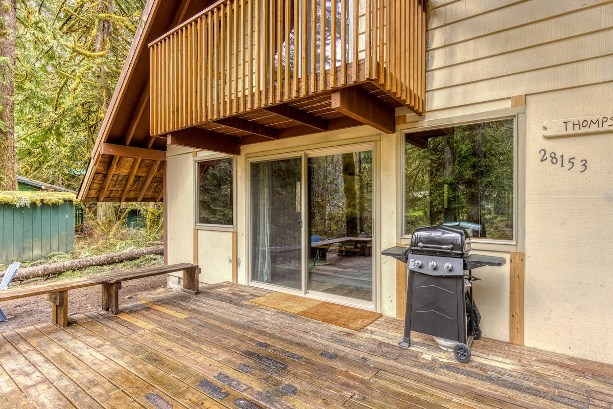 The back exterior of the cabin featuring a spacious wooden deck with a gas BBQ grill, a sliding glass door, and a small wooden bench, all surrounded by lush green trees.