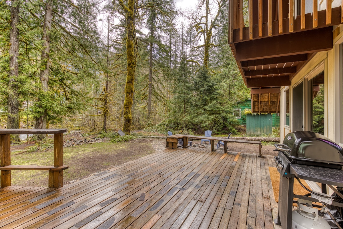 A wide view of a large wooden deck extending from the cabin, equipped with a BBQ grill and a wooden table, overlooking a natural landscape of tall evergreen trees and a hint of a river or stream in the distance.