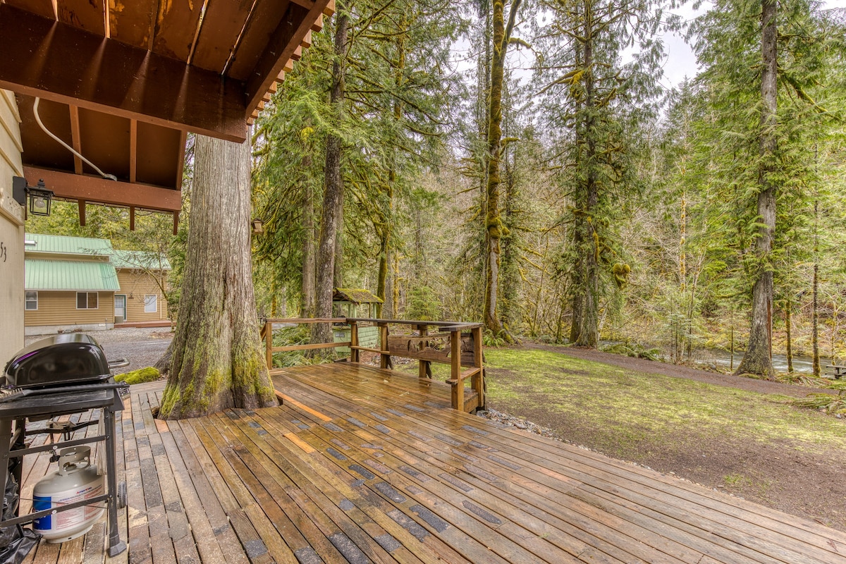 A wooden deck with a BBQ grill and a propane tank, extending into a natural setting with tall trees and a visible river or stream flowing in the background, highlighting the riverfront aspect of the property.