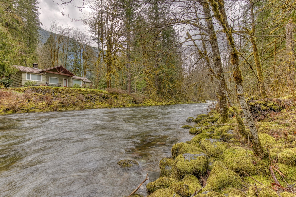 A scenic river with moss-covered rocks in the foreground, flowing past a distant cabin nestled on a raised, tree-lined bank under a cloudy sky.