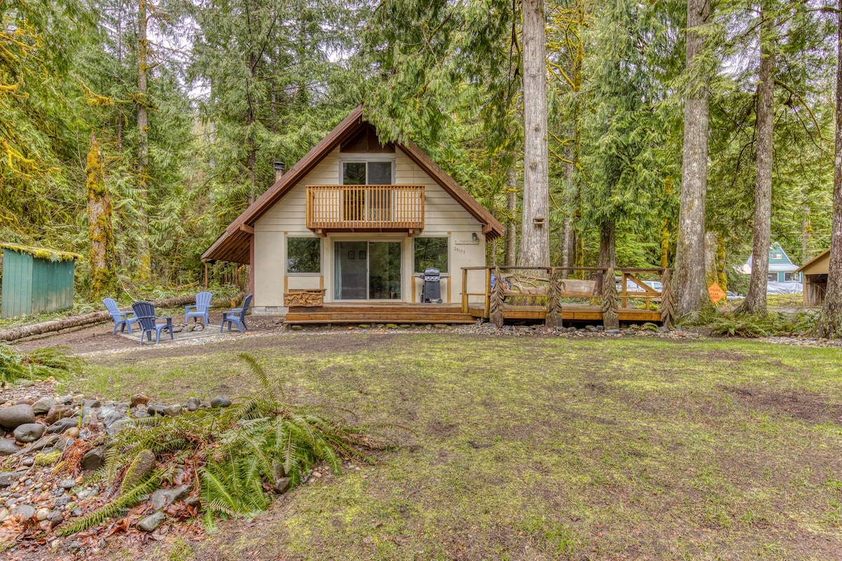 A-frame cabin with light-colored siding and a wooden balcony, surrounded by dense evergreen trees and a grassy yard with a fire pit area and blue Adirondack chairs.