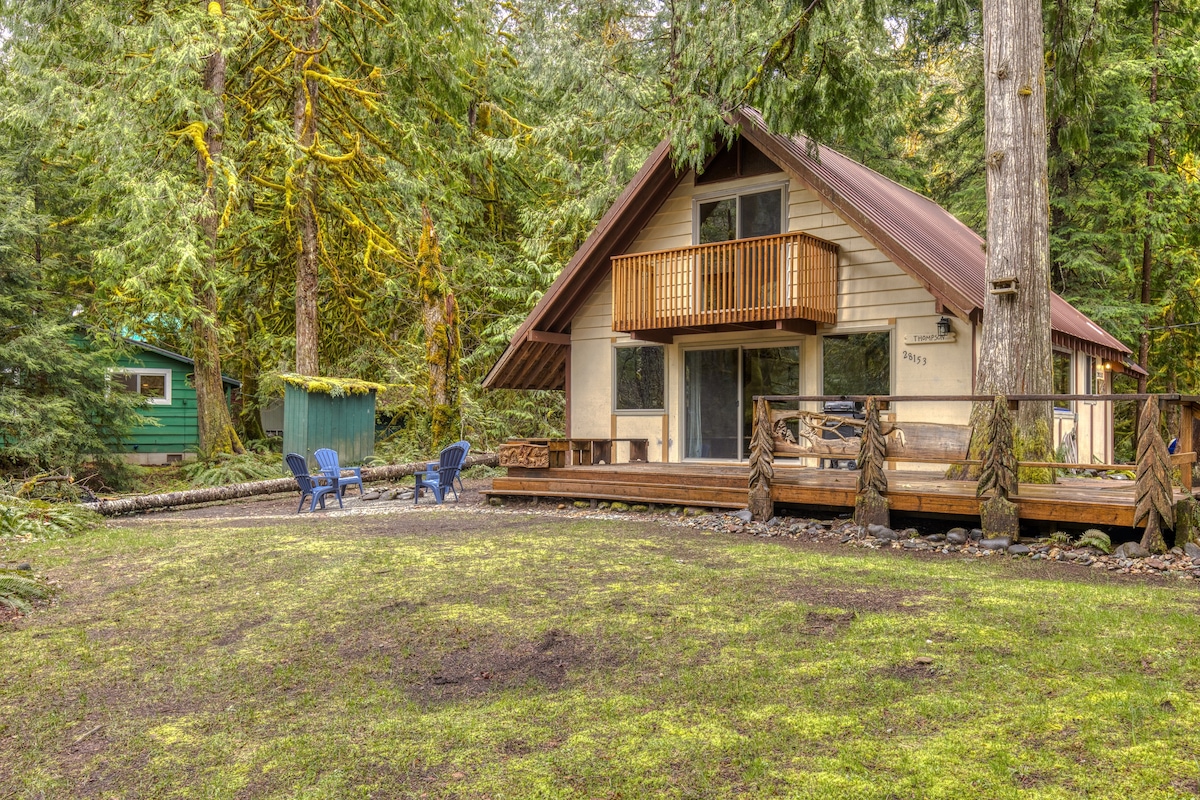 A-frame cabin with light-colored siding and a brown roof, featuring a wooden deck, a second-story balcony, and surrounded by lush green trees and mossy ground.