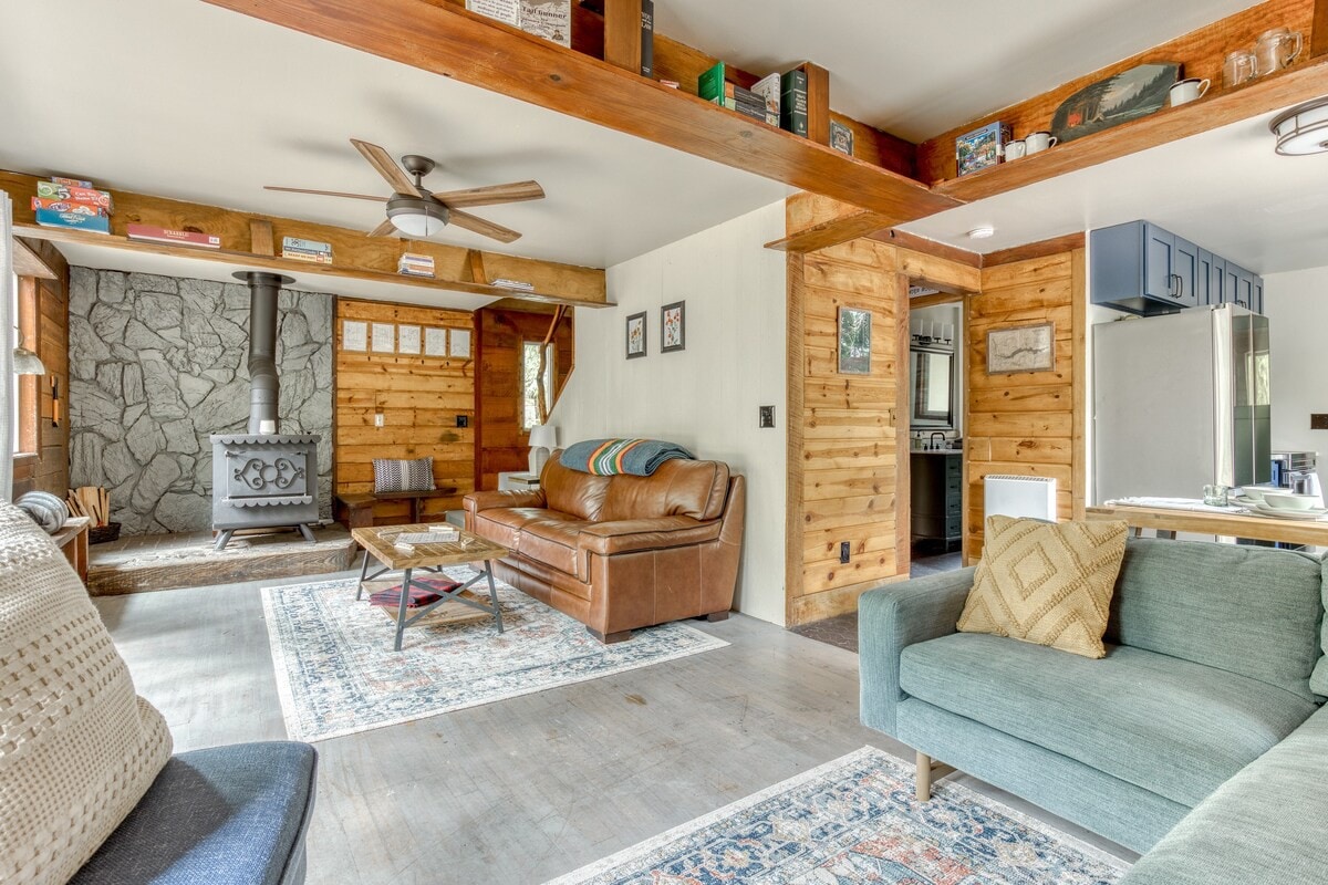 Inviting living room featuring a prominent wood-burning stove set against a stone accent wall, with a brown leather sofa and a light blue sectional on a patterned rug, under exposed wooden beams.