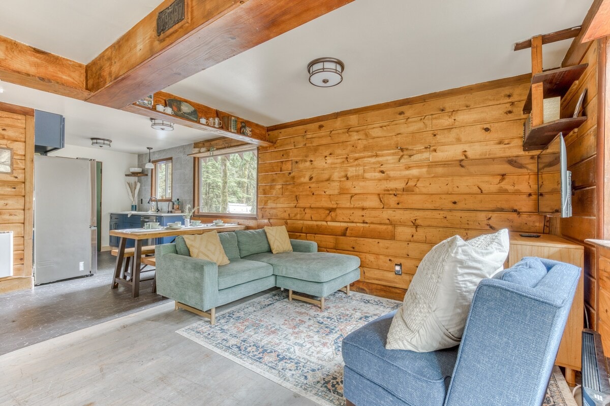 Open-concept living space with a light blue sectional sofa on a patterned rug, transitioning into a dining area with a wooden table and a modern kitchen with blue cabinets and a refrigerator.