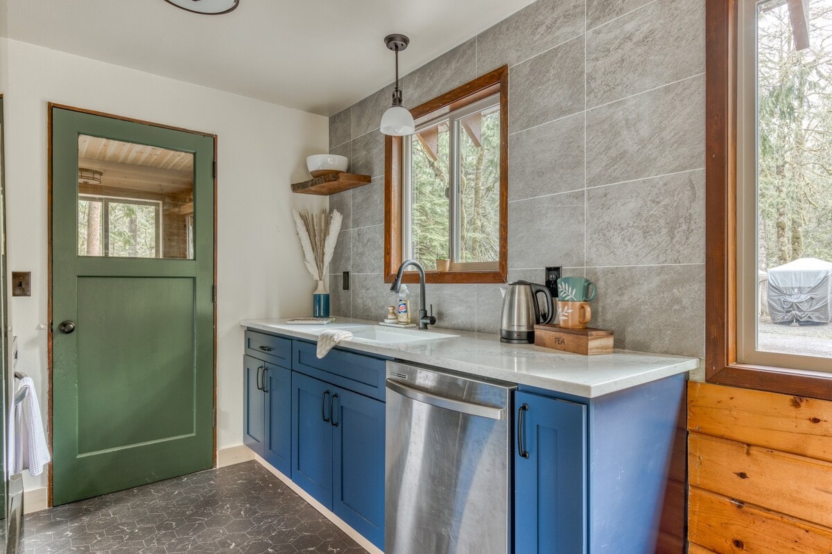 A bright kitchen area featuring blue base cabinets, white countertops, a stainless steel dishwasher, and a black faucet over a white sink, with a green door and large window.