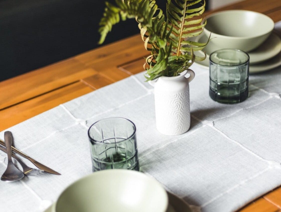 Close-up of a dining table set with light green bowls and plates, dark green glasses, and a white vase with a fern, all on a white linen runner.