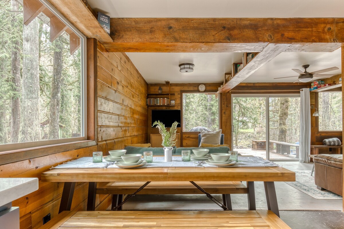 Spacious dining area with a long wooden table and benches, set against a backdrop of large windows offering forest views, and opening into a living room with a sofa.