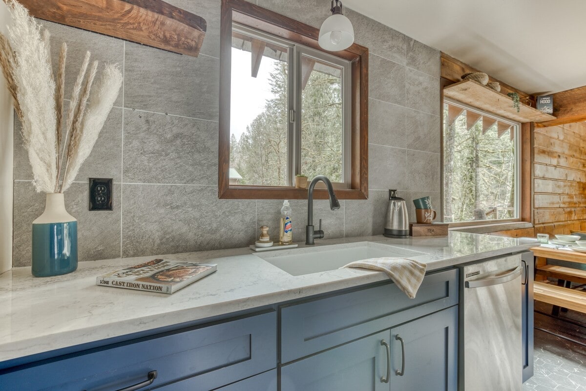 Modern kitchen with blue base cabinets, light gray quartz countertops, and a gray tiled backsplash. A large window above the sink offers a view of trees, and a white vase with pampas grass adds a decorative touch.